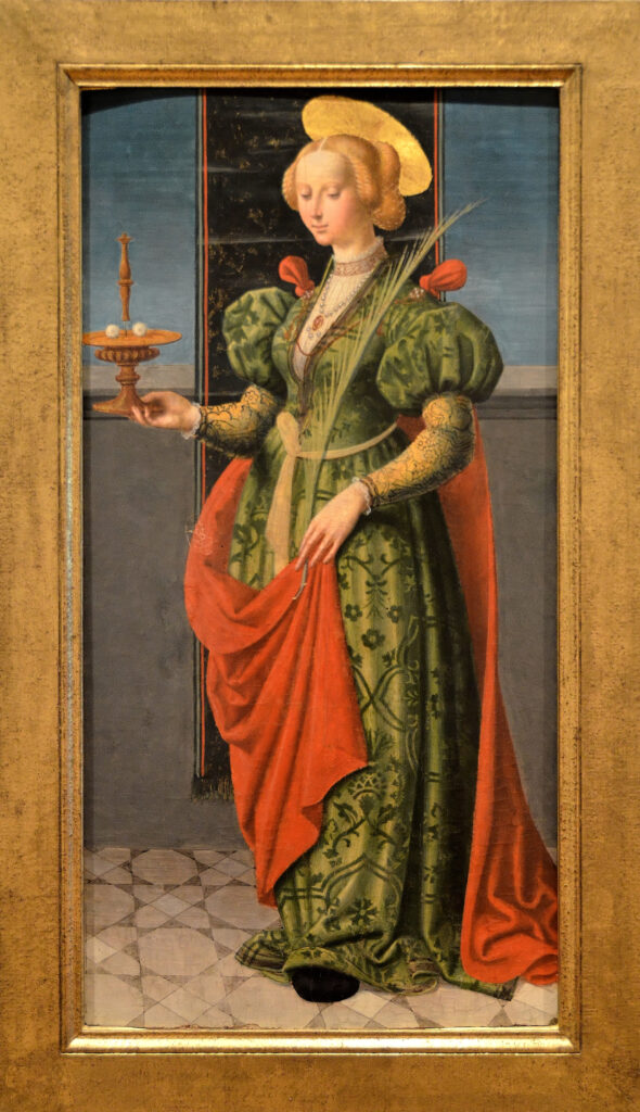 Early 16th-century Painting of St Lucy wearing a green robe with a red cloak, and halo. In her right hand, she holds a lamp with a pair of eyeballs resting on it--both the lamp and the eyes are attributes of St Lucy as patron saint of blind people, with reference to medieval legends about her martyrdom; painting in the Musee des Beaux Arts, Nancy (Meurthe-et-Moselle), attributed to Pseudo-Giovenone (around 1510); via Wikimedia Commons