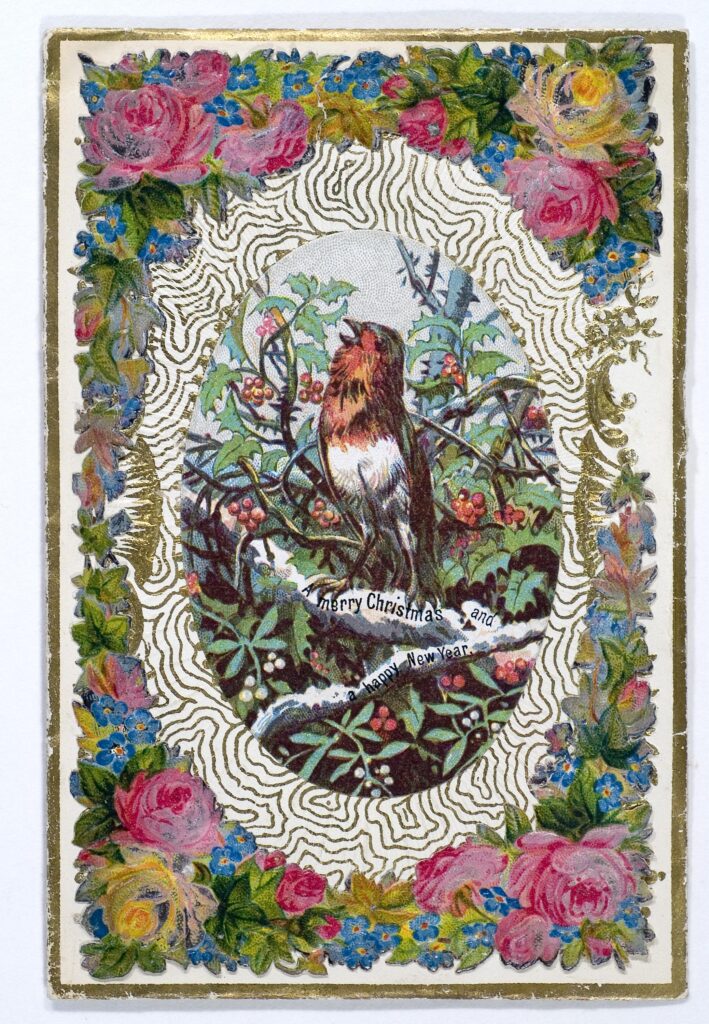 Victorian Christmas card in The Folklore Society Archives, showing a singing robin on a branch surrounded by greenery, in a white oval with a lace effect, and bordered with pink flowers and greenery