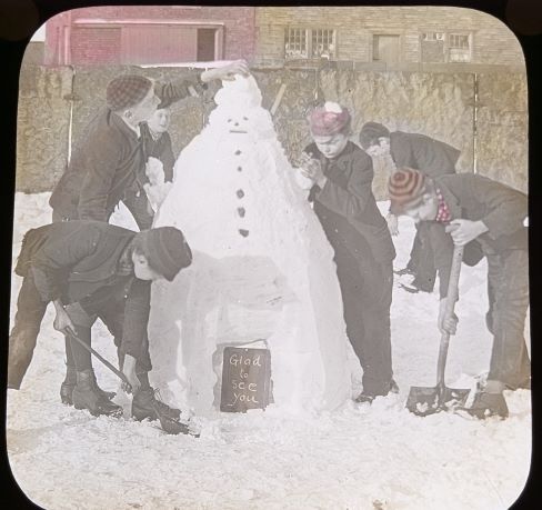 Hand-coloured photograph magic lantern slide from around the 1910s showing a group of 6 boys making a large snowman, with shovels and hands. The snowman has coals or stones for face and buttons, and at its base is a school slate with the chalked message 'Glad to See You.' Digital photo Nigel Longstaff.